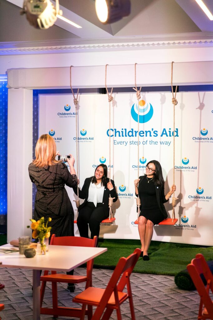 Two women sitting on swings in front of a step and repeat