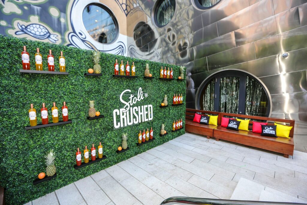 Large greenery wall decorated with bottles of alcohol and pineapples a logo that says "Stoli Crushed"
