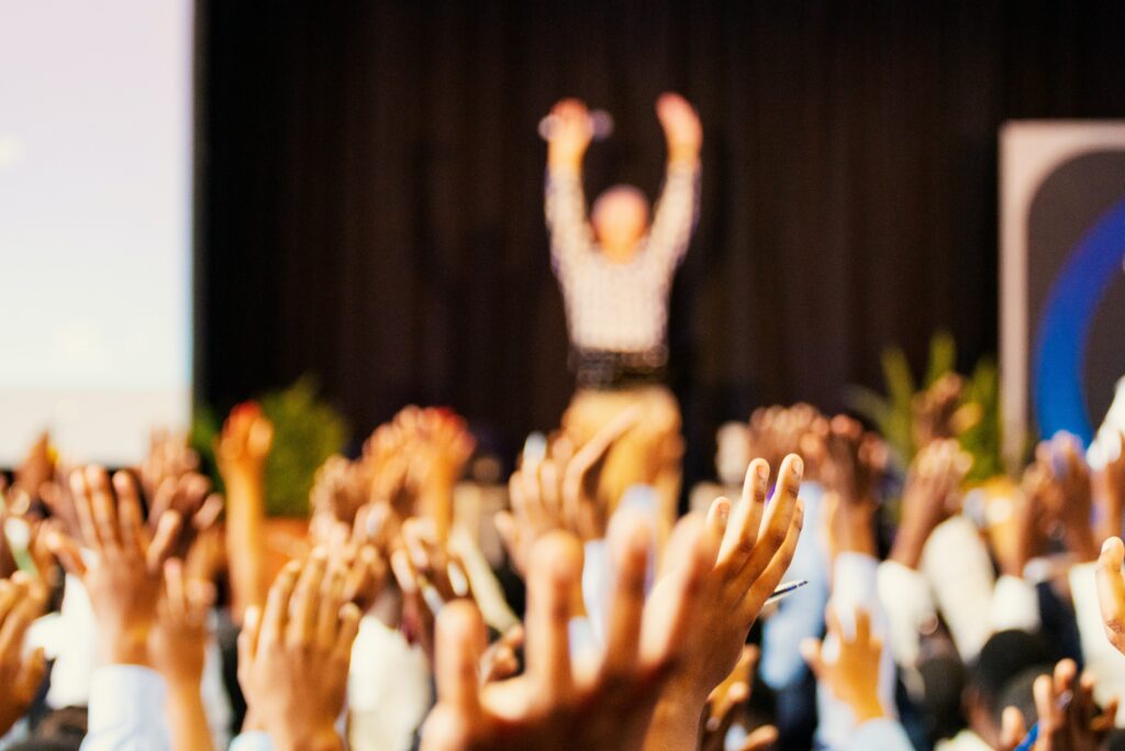 A person on stage with their hands raised in the air in front of a large group of people with their hands raised in the