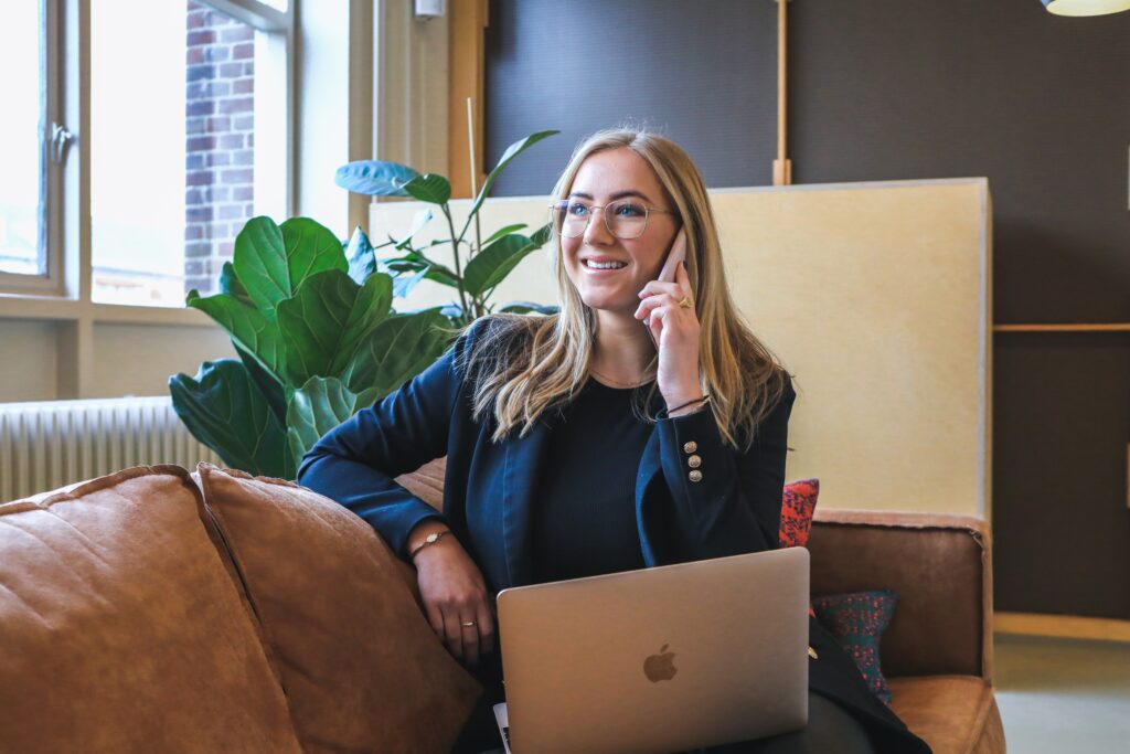 A blonde woman in a blazer and glasses on her phone and computer 