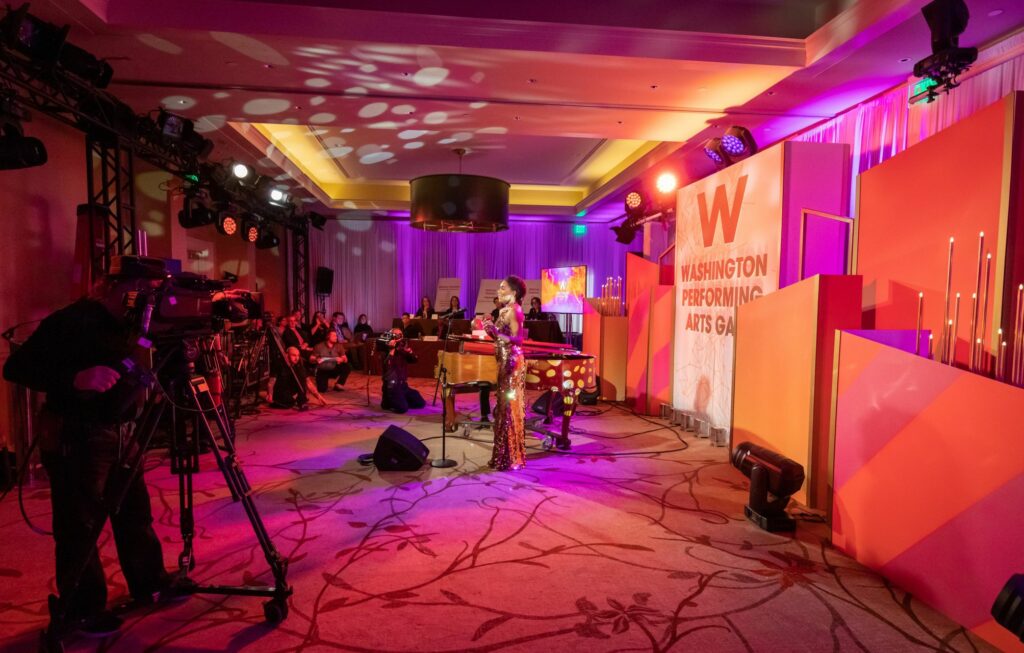 A woman standing at a microphone in front of a backdrop with camera equipment filming her