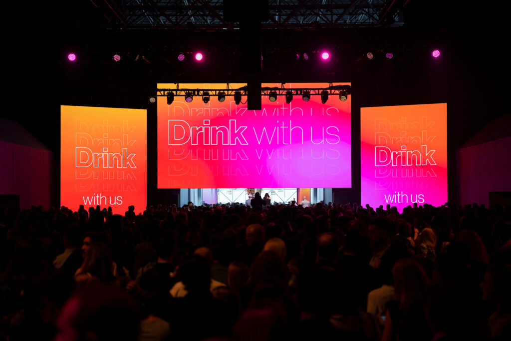 Stage at an event with large screens that say "Drink with us"