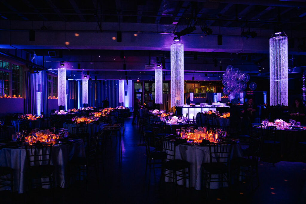 Large ballroom with dark purple lights and candles on the tables and long white cylinder light fixtures