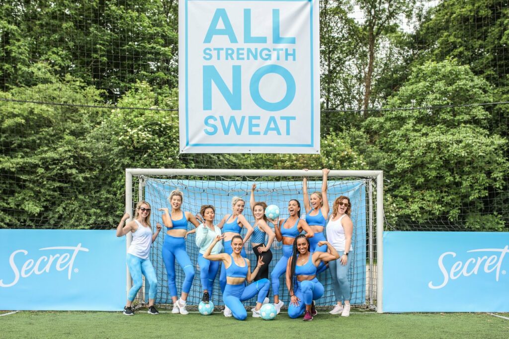 Women standing in front of soccer goal with sign of above them that reads, 'All Strength No Sweat'