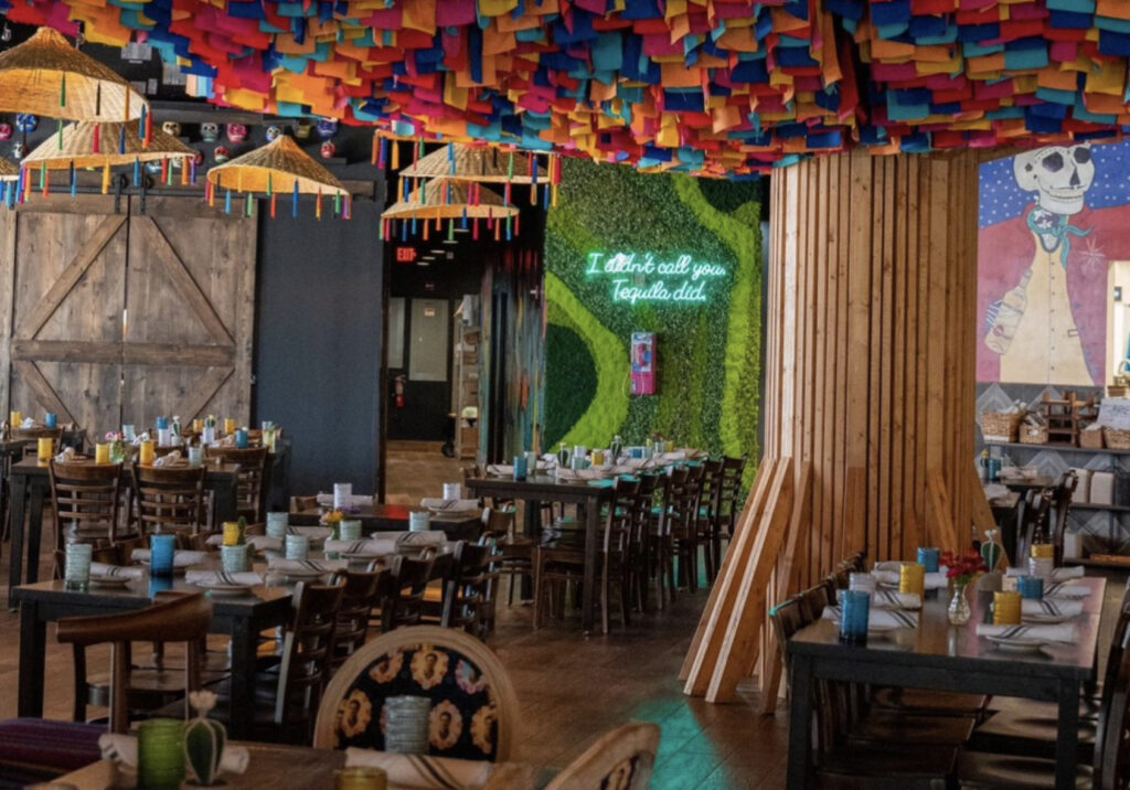 Kavas Tacos & Tequila restaurant tables and chairs with green living backdrop wall and neon signage and colorful tree ceiling. 