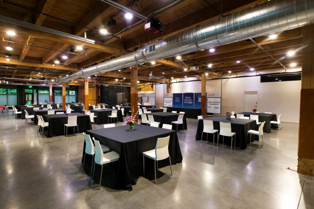 A spacious indoor venue with dominant colors of brown and grey, with accents of black. The tables and chairs are arranged in a neat and orderly fashion. The ceiling is high and features several light fixtures, providing ample lighting for the space.