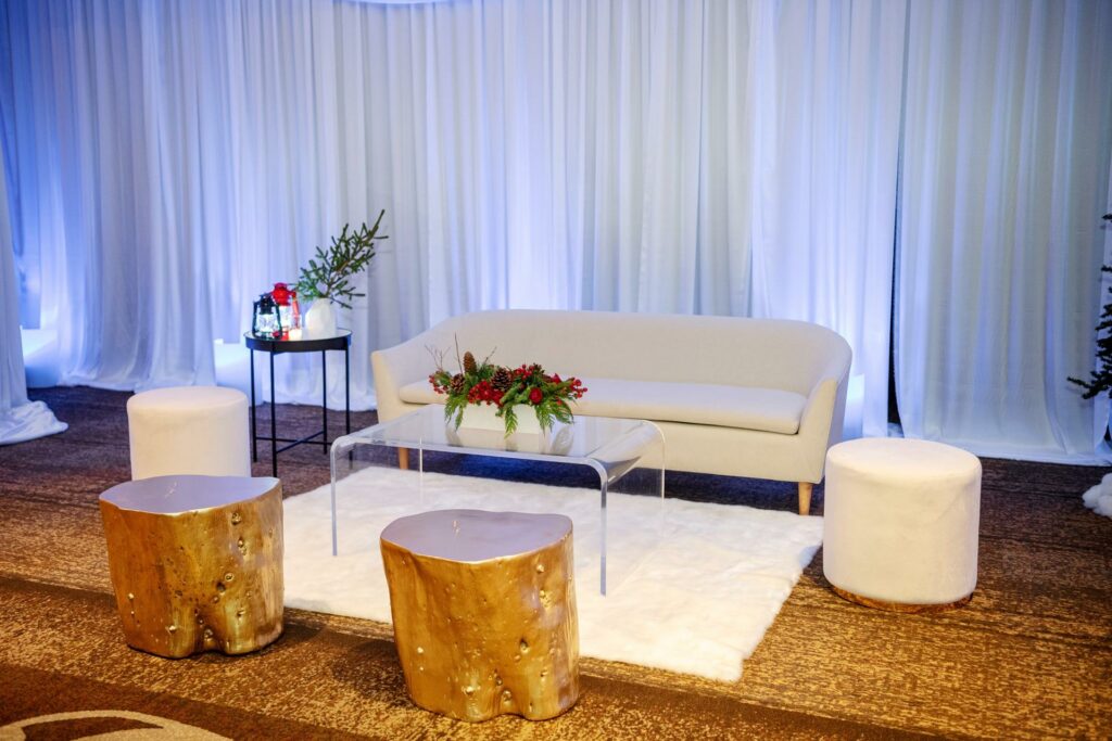 A beautifully decorated room with white couch, a table adorned with flowers, two cylindrical-shaped stools in crisp white, two wooden stools painted in gold, and gold carpet covering the floor.