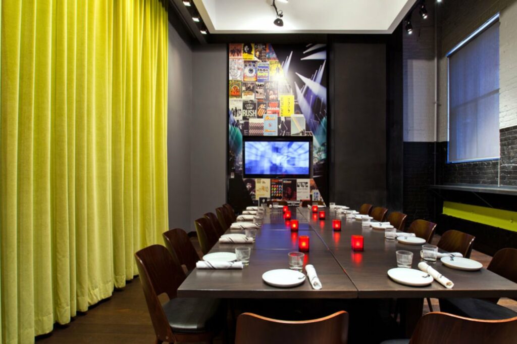 A private dining room with a curtain on the other side and an exposed brick wall on the other, a long table in the middle and an LCD TV screen