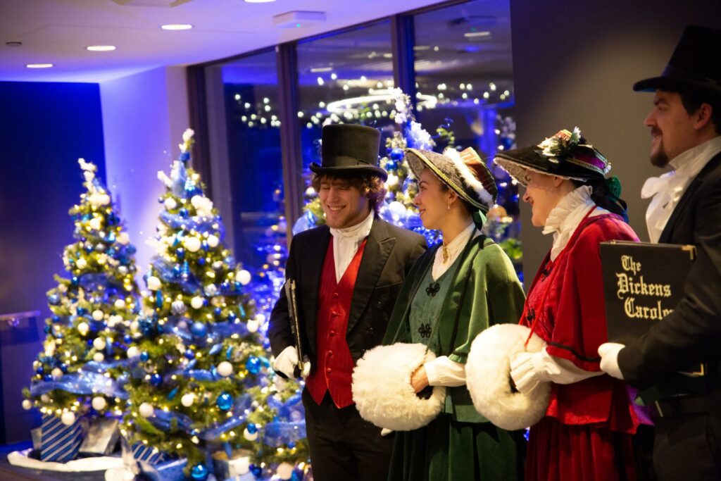A room with four people standing on the right wearing holiday themed costumes, and two big Christmas trees on the left.