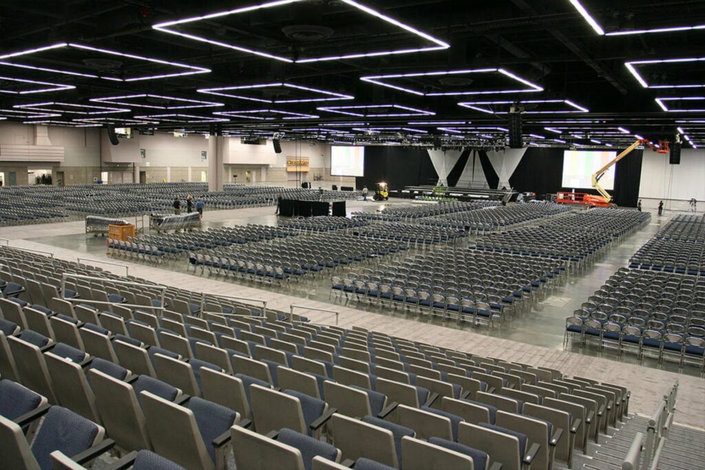 A huge convention hall with plenty of fixed chairs, exposed ceiling and a stage