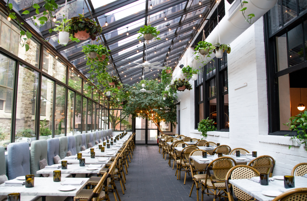A bright glass-enclosed patio and hanging plants and cozy tables and chairs 