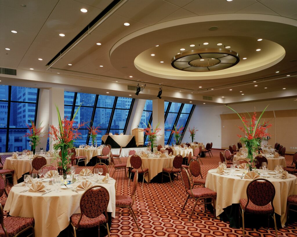 A huge ballroom with floor to ceiling windows overlooking the views of the city and plenty of table and chairs 
