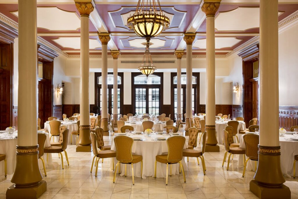 A unique indoor room adorned with a color palette of gold and white, an elegant chandelier at the center, and large windows at the back.  