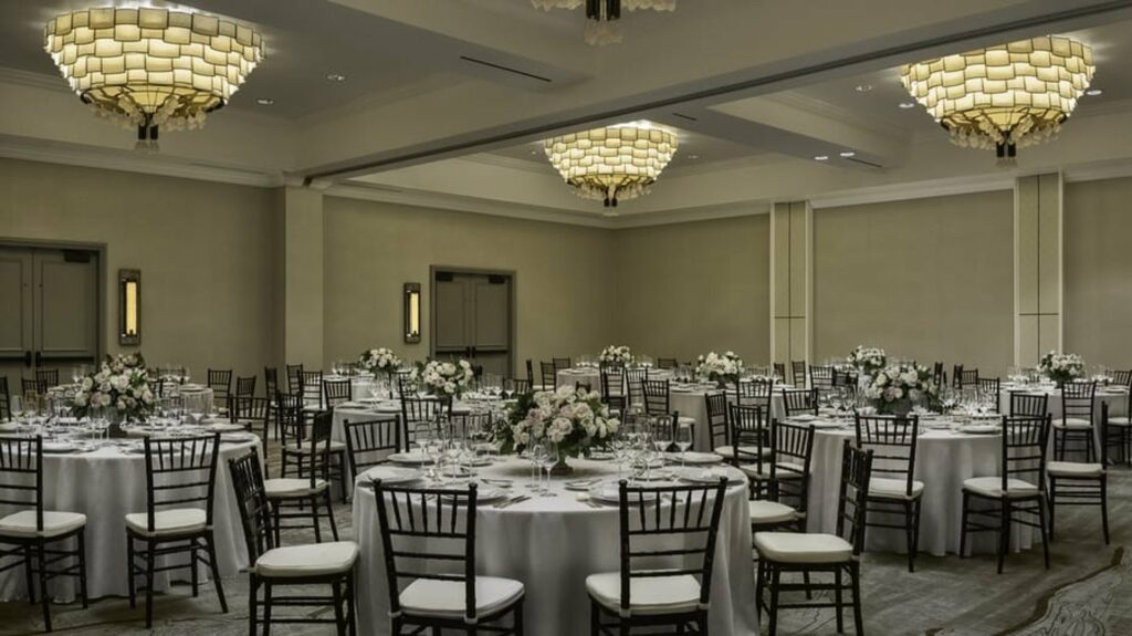 An elegant indoor space with three beautiful chandeliers and white-cloth tables and chairs around it. 