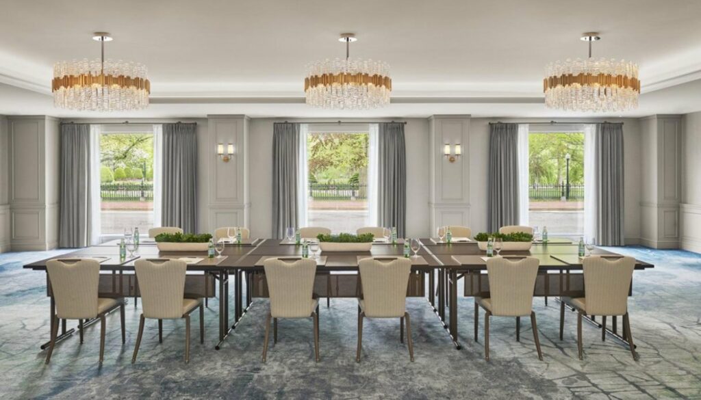 An elegant meeting room with overlooking view from the floor-to-ceiling windows, tables and chairs arranged at the center, and three beautiful chandeliers hanging above it. 