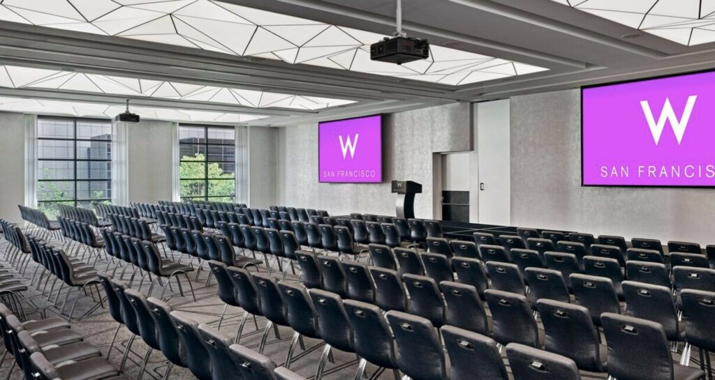 A large event space with floor to ceiling windows, 2 projector screens, and plenty of chairs in theatre style setup