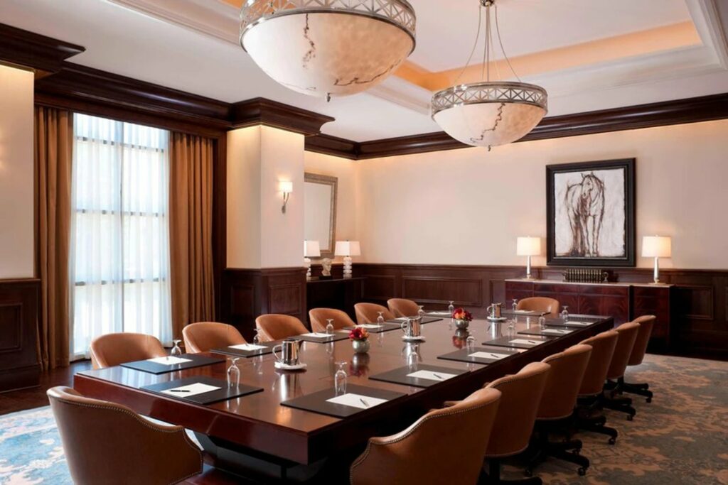 An elegant-looking meeting room with 2 big lighting fixtures,  a conference table in the middle and a floor to ceiling windows offering natural lighting.