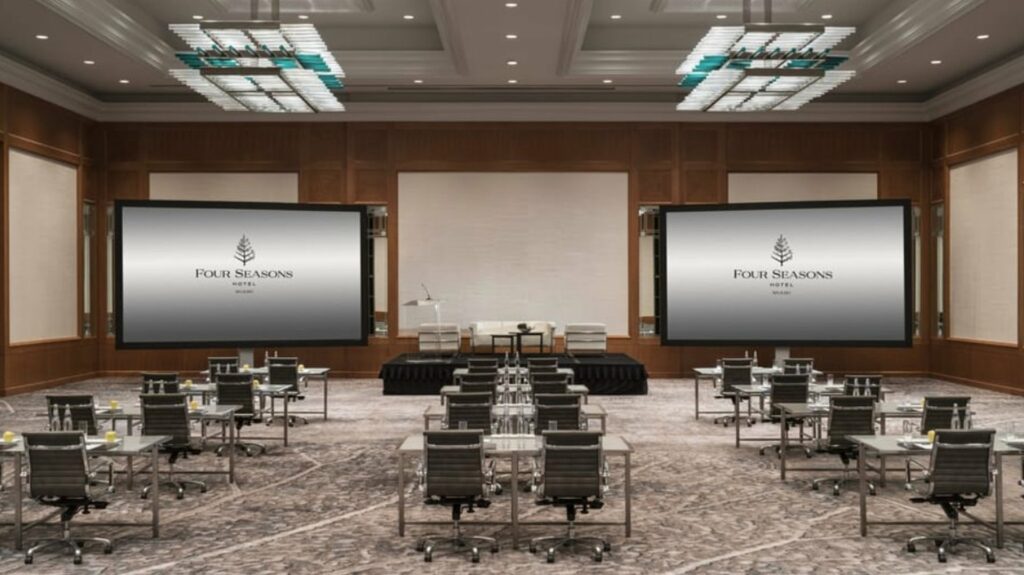 A spacious room with high ceiling, tables and chairs arranged neatly, and 2 big screens at the front. 