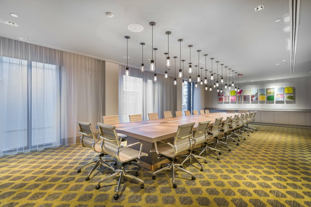 An elegant meeting room with natural light that floods in through floor-to-ceiling windows covered with thin white curtain, with a long table at the center and chairs around it. 
