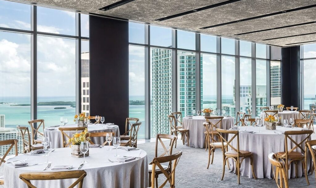 An elegant room offering panoramic views of a cityscape and waterfront through floor-to-ceiling windows, with tables and wooden chairs around it. 
