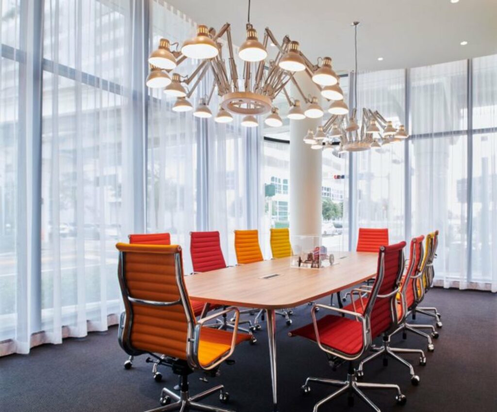 A meeting room with elegant chandelier, a long table at the center with vibrant colored chairs around it and an overlooking view from the outside through floor-to-ceiling windows. 
