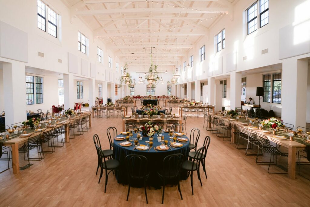 An elegant and spacious room with natural light streams through the large windows, high ceiling adorned with string lights, along with the warm wooden flooring, and careful arranged tables with decorations and chairs. 