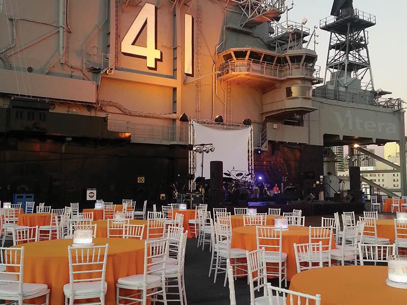 An outdoor event place on board a large ship docked at a pier with tables and chairs arranged. 